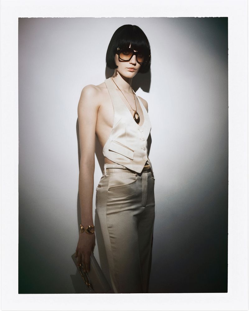 Fashion shot of a woman with bobbed hair wearing sunglasses, a cream-colored halter top and pants, and gold jewelry in a studio.