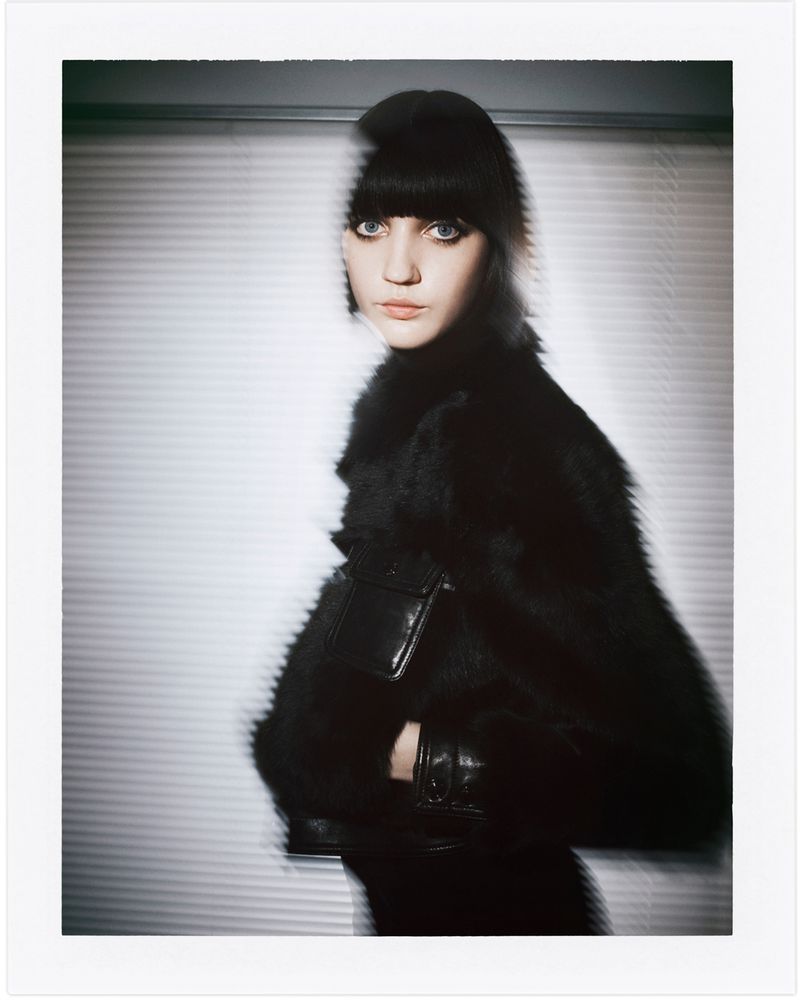 Fashion shot of a young woman in a black fur coat with a fringe, against a backdrop of white Venetian blinds in soft studio lighting.