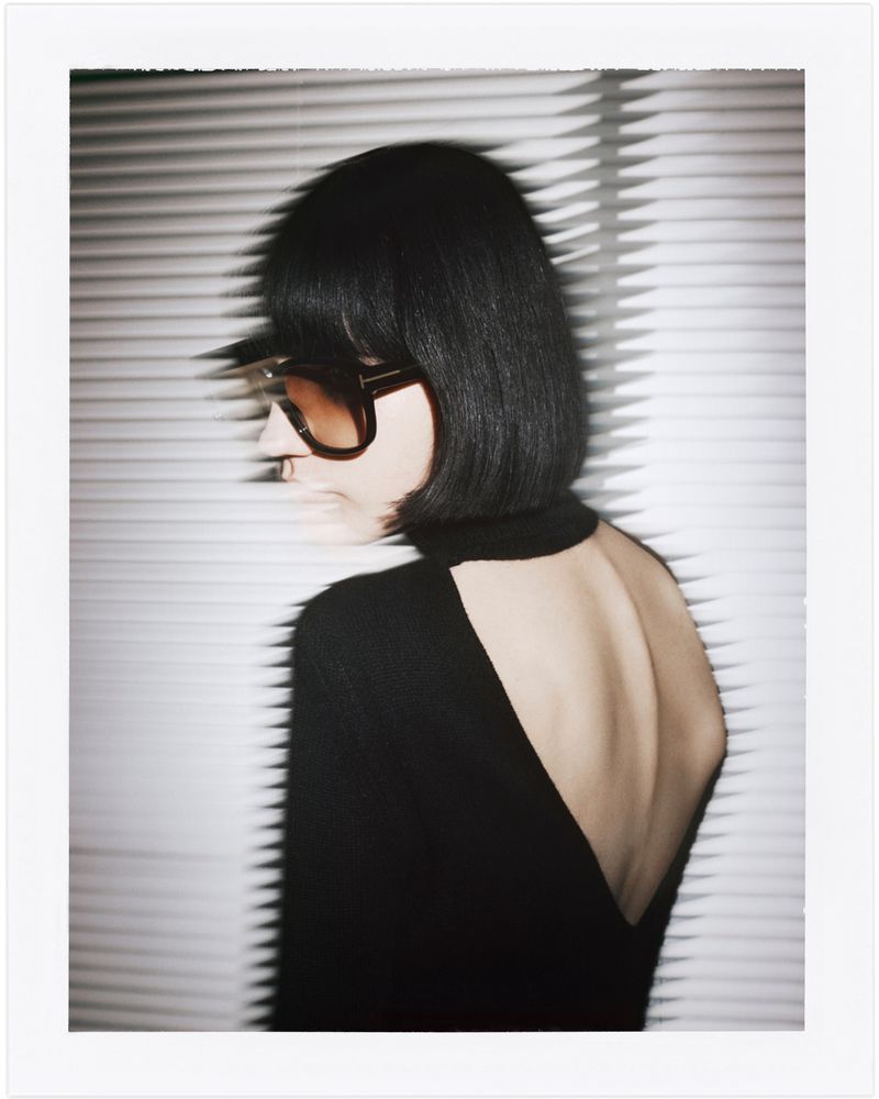 Profile of an adult female with sunglasses wearing a black turtle neck dress with an open back in front of a window with blinds.