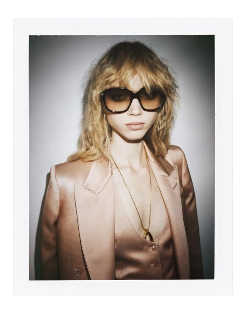 A blonde adult woman poses wearing sunglasses, blazer, blouse, and pendant necklace against a grey-white background.