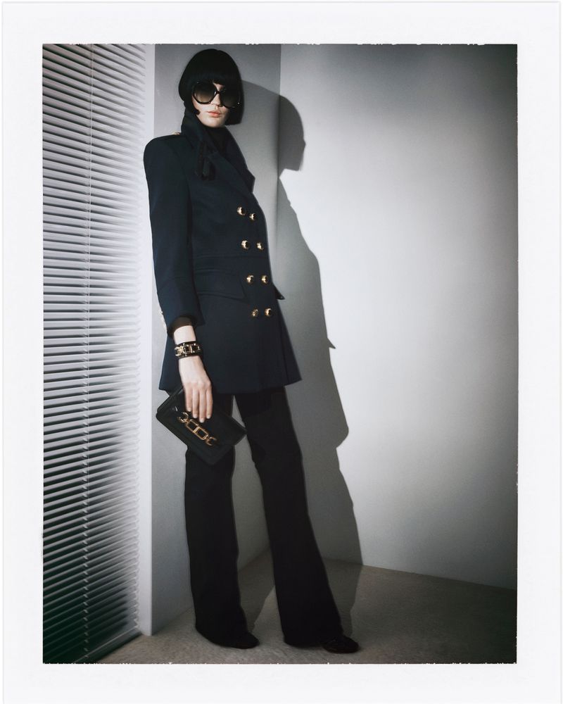 Woman in a double-breasted black blazer, black pants, and sunglasses poses in front of a wall with window blinds and her shadow.