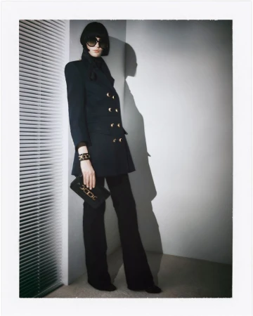 Woman in a double-breasted black blazer, black pants, and sunglasses poses in front of a wall with window blinds and her shadow.