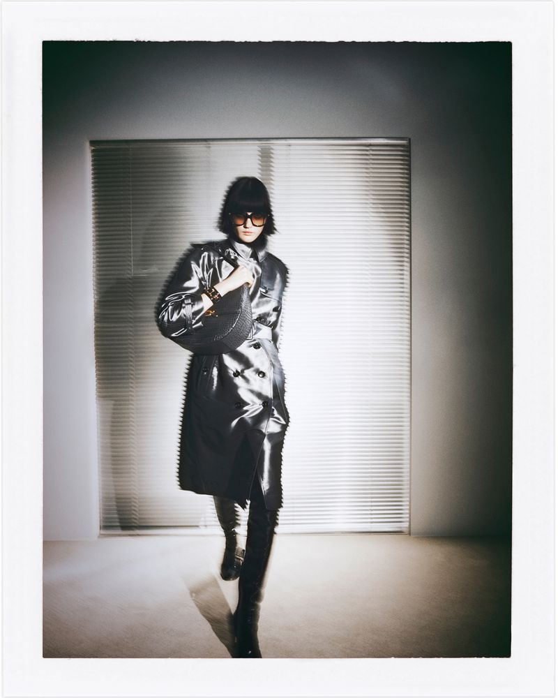 A single adult stands in a photo studio wearing a silver trench coat and dark sunglasses while carrying a matching silver bag.
