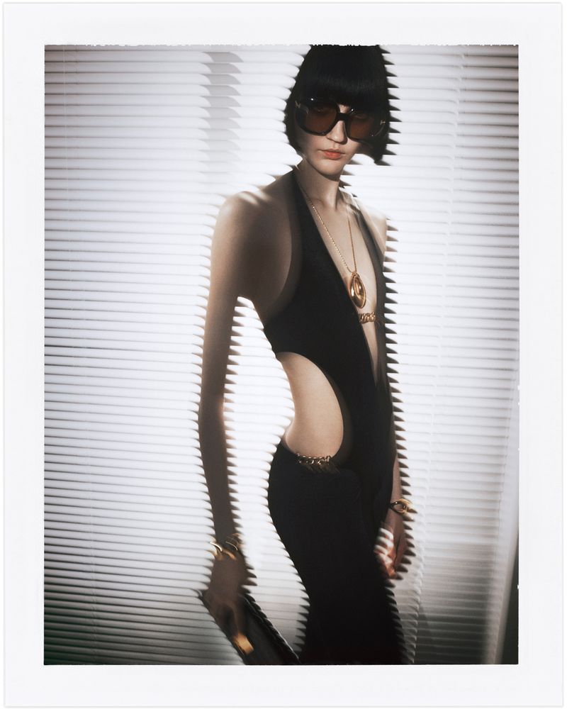 Fashion photo of a woman with black hair and sunglasses wearing a black dress with cutouts, against a white-louvered background.