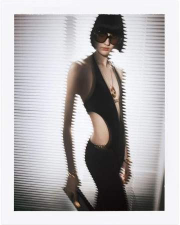 Fashion photo of a woman with black hair and sunglasses wearing a black dress with cutouts, against a white-louvered background.