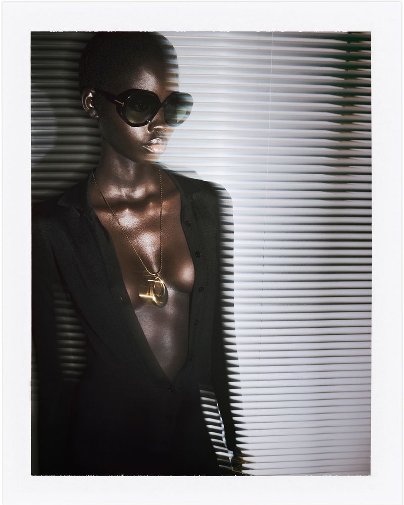 Model wearing black oversized sunglasses, an open black shirt, and a gold pendant against slatted venetian blinds.