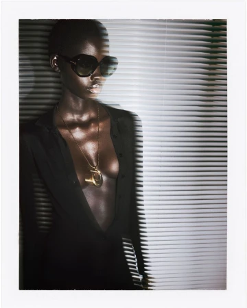 Model wearing black oversized sunglasses, an open black shirt, and a gold pendant against slatted venetian blinds.