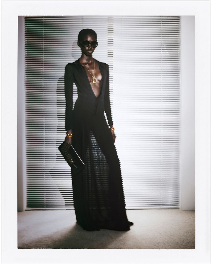 Elegant African-American woman in black gown, sunglasses, and gold jewelry poses in a light-filled studio near venetian blinds.