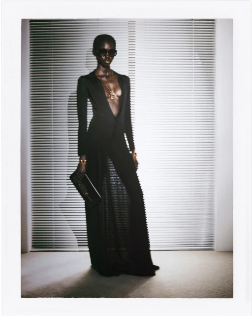 Elegant African-American woman in black gown, sunglasses, and gold jewelry poses in a light-filled studio near venetian blinds.