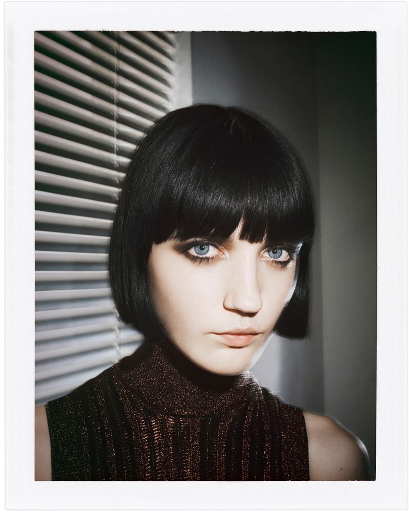 Close-up portrait of an adult female with a dark bob haircut, wearing a high-neck top, and posing in front of blinds, beauty shot.