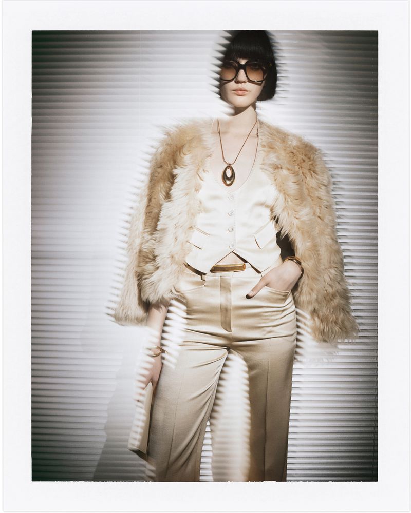 Fashion shot of a female model wearing a fur coat, glasses, necklace, and light-colored silk suit, posing in front of a striped background.