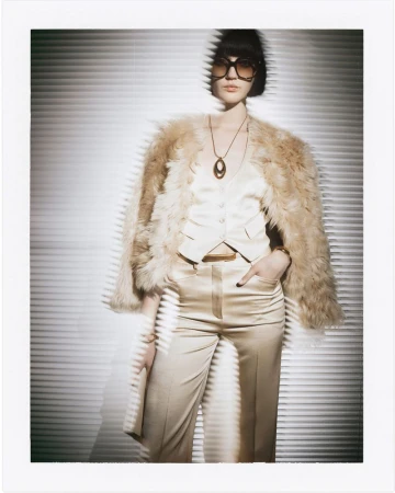 Fashion shot of a female model wearing a fur coat, glasses, necklace, and light-colored silk suit, posing in front of a striped background.