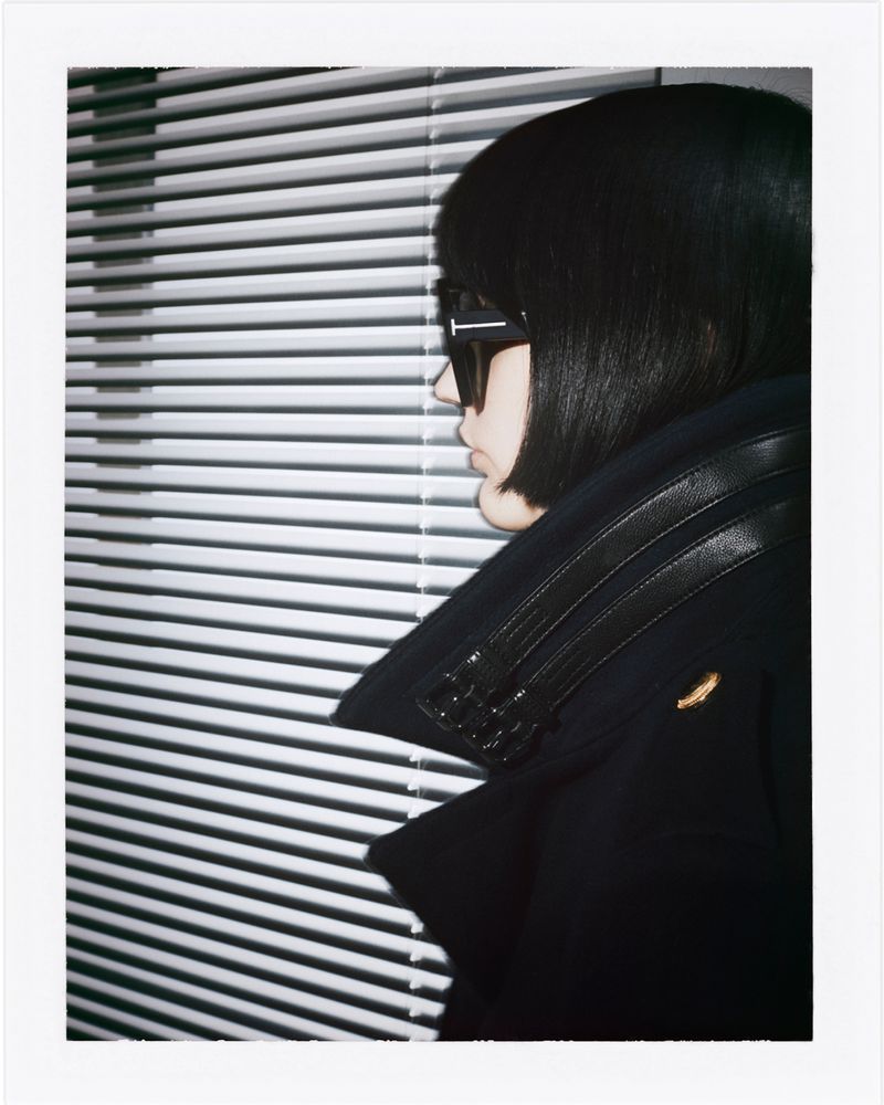 Fashion shot of a woman in sunglasses and a dark jacket posing in front of window blinds. The shot is portrait-oriented.