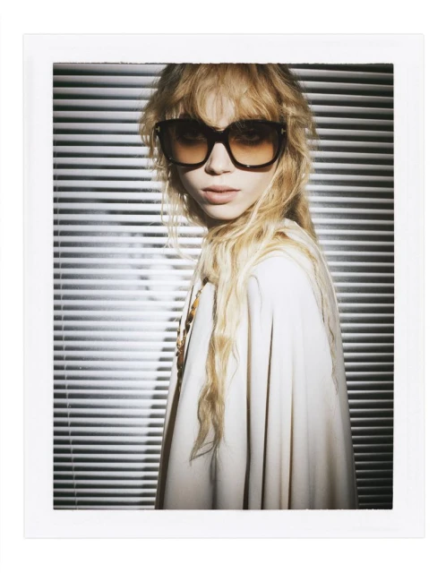 Portrait of a blonde woman with sunglasses in front of window blinds, fashion editorial style. Creamy light and Polaroid frame.