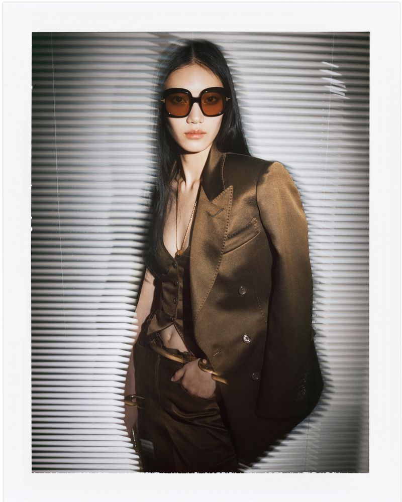 Elegant Asian woman in a bronze suit, sunglasses, and gold bracelets poses against a white Venetian blind background in a fashion shot.