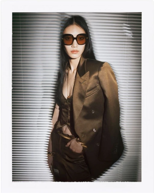Elegant Asian woman in a bronze suit, sunglasses, and gold bracelets poses against a white Venetian blind background in a fashion shot.