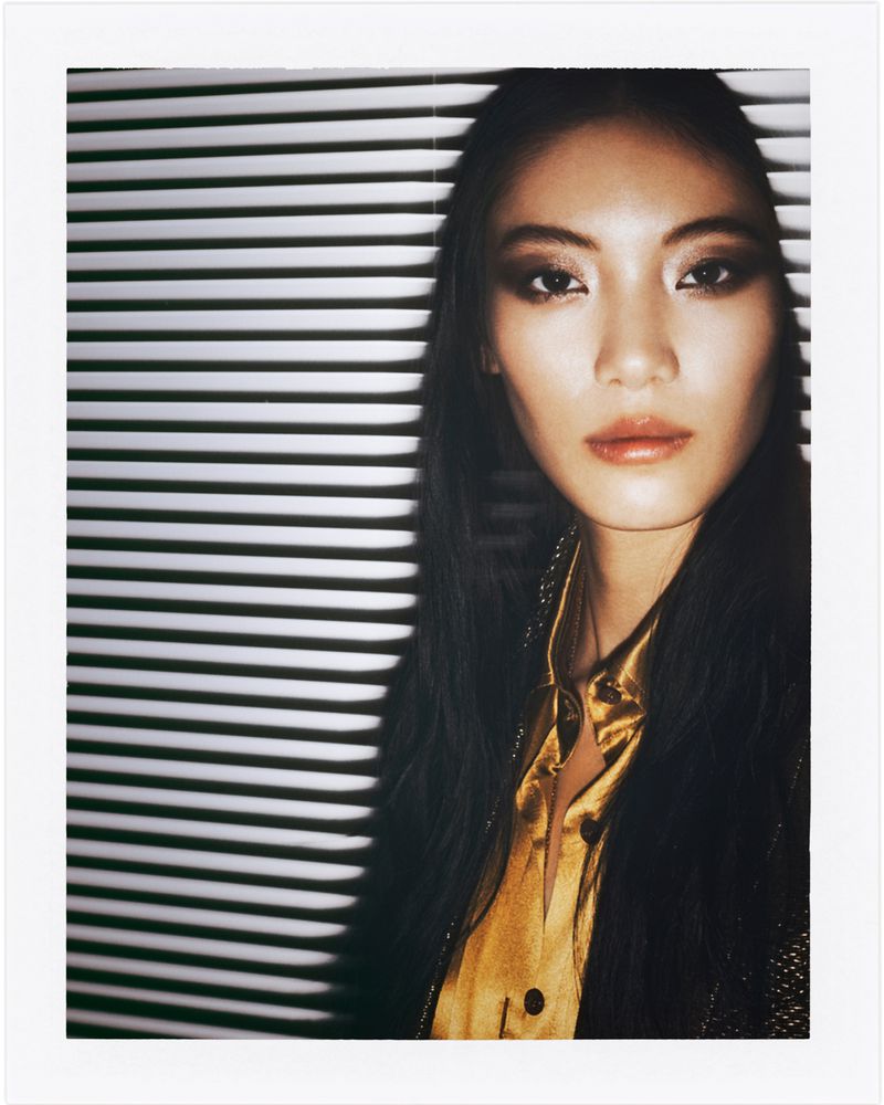 Portrait of an Asian woman with long black hair, wearing a gold blouse, posing in front of a background of horizontal blinds.