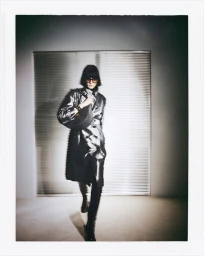 Model wearing a glossy metallic coat and sunglasses holding a dark handbag against a window with blinds