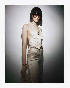 Model wearing a champagne satin halter waistcoat, matching trousers, sunglasses, and gold jewelry against a studio backdrop.