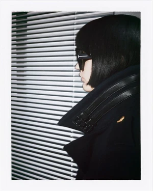 Model in profile wearing black sunglasses and a dark coat against horizontal window blinds