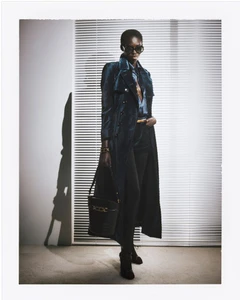 Model wearing dark sunglasses and a long dark blue coat holding a black handbag against window blinds.