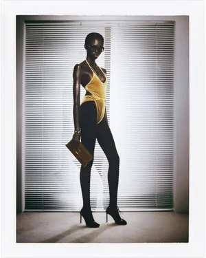 Model wearing a yellow halter bodysuit and black leggings holding a clutch in front of window blinds