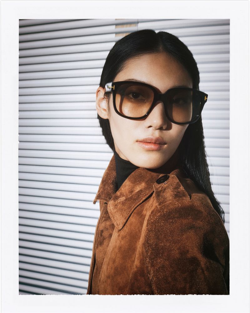 Model wearing dark sunglasses and brown suede jacket against horizontal window blinds