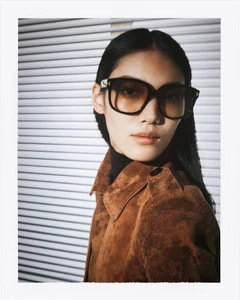 Model wearing dark sunglasses and brown suede jacket against horizontal window blinds