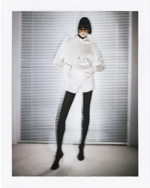 Model wearing dark sunglasses and a white hooded sweatshirt standing against horizontal window blinds.