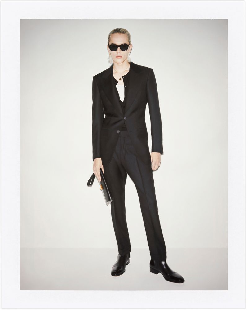 Adult in a dark suit with black sunglasses poses in a photo studio against a white background. Clutch in hand and dark shoes.