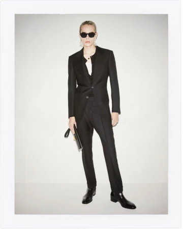 Adult in a dark suit with black sunglasses poses in a photo studio against a white background. Clutch in hand and dark shoes.