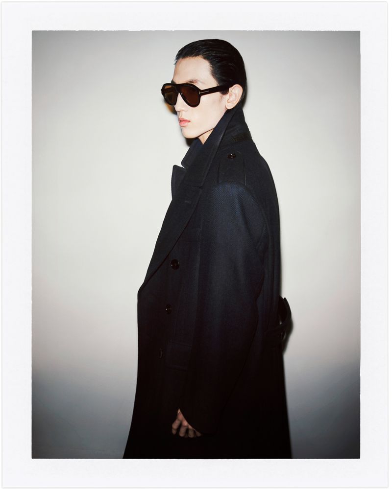 A man in a dark overcoat and sunglasses poses against a white backdrop, with a side view highlighting his angular features and fashion attire.