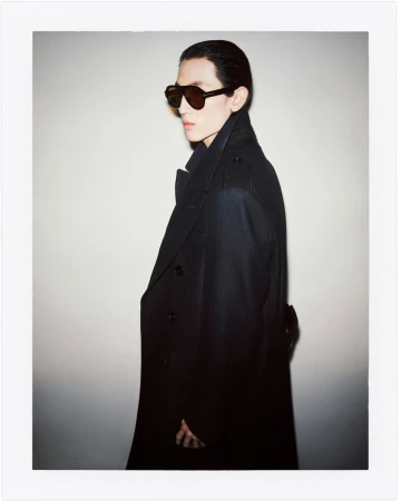 A man in a dark overcoat and sunglasses poses against a white backdrop, with a side view highlighting his angular features and fashion attire.