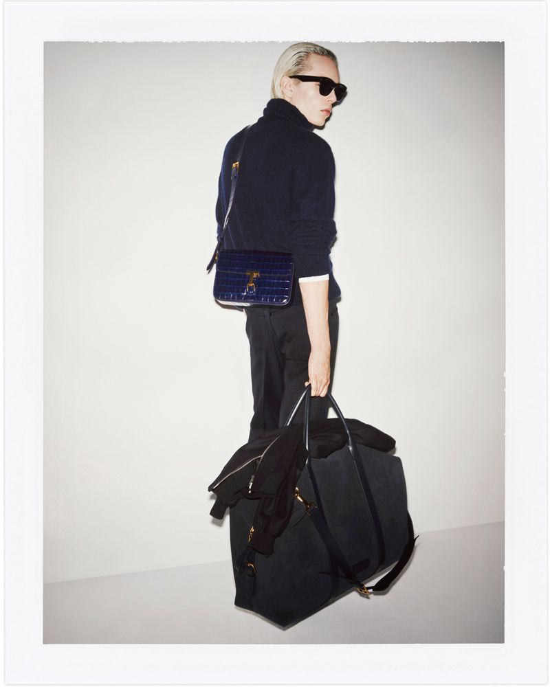 Stylish man with platinum blond hair wearing a dark turtleneck and sunglasses, posing with a navy blue bag and duffel bag against white.