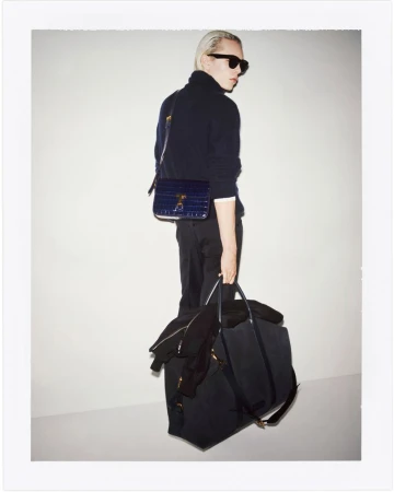 Stylish man with platinum blond hair wearing a dark turtleneck and sunglasses, posing with a navy blue bag and duffel bag against white.
