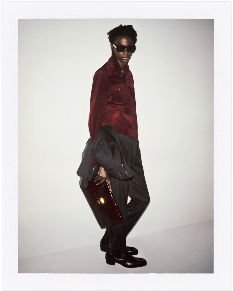A stylish black man with sunglasses poses in a photo studio wearing a red corduroy shirt, grey suit jacket, trousers, and black leather shoes.