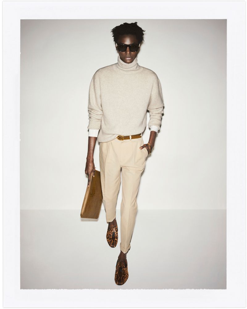 Full-length shot of a black male model in beige turtleneck, pants, animal print shoes, holding a clutch, posing in a photo studio.