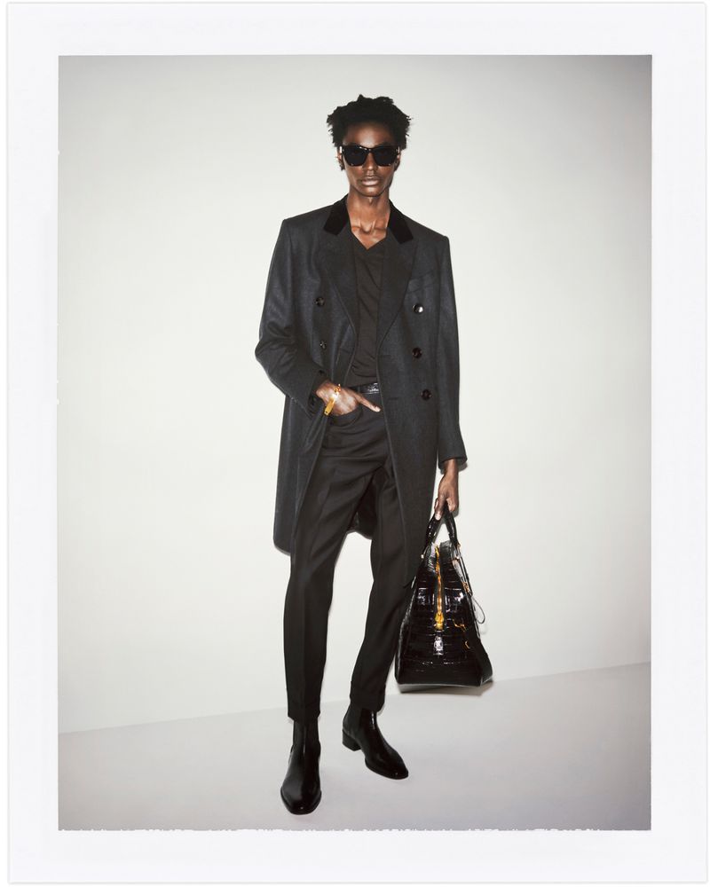 Stylish Black male fashion model poses in a coat, dress pants, sunglasses, and ankle boots holding a black leather duffel bag.