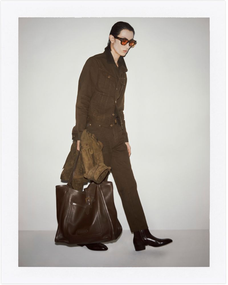 A person wearing a brown jacket and pants, carrying a brown leather tote bag and jacket, poses in a studio with a white backdrop.