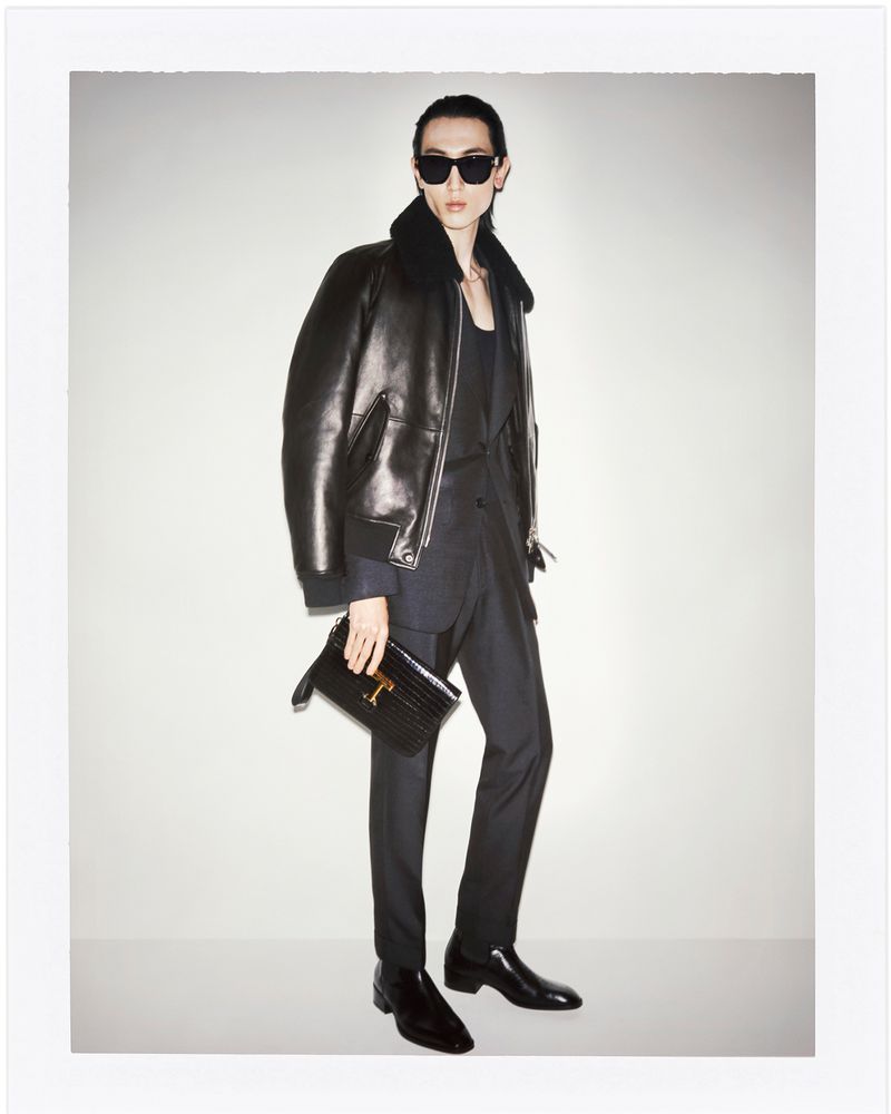 A stylish individual wearing a black leather jacket, dark suit, sunglasses, and shiny boots, carrying a black clutch against a white backdrop.