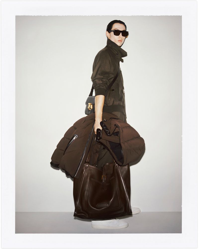A stylish adult in sunglasses stands indoors in a jacket, holding a brown puffer coat and large leather bag in a fashion setting.