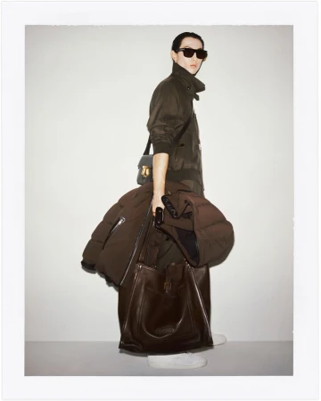 A stylish adult in sunglasses stands indoors in a jacket, holding a brown puffer coat and large leather bag in a fashion setting.