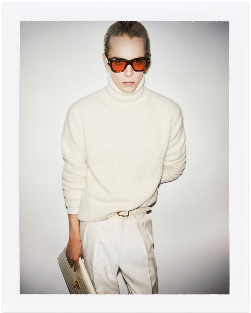 Man in a turtleneck sweater and orange sunglasses, holding a document, poses against a white background in a studio-style setting.
