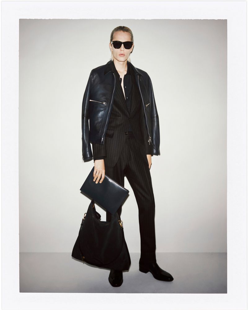 Fashionable adult woman wearing a leather jacket and pinstripe suit, posing indoors with a large handbag and document case.