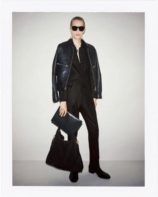 Fashionable adult woman wearing a leather jacket and pinstripe suit, posing indoors with a large handbag and document case.