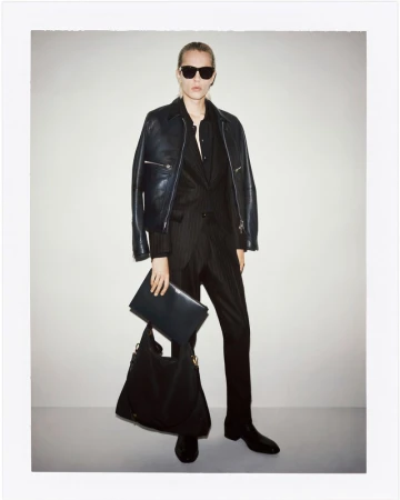 Fashionable adult woman wearing a leather jacket and pinstripe suit, posing indoors with a large handbag and document case.