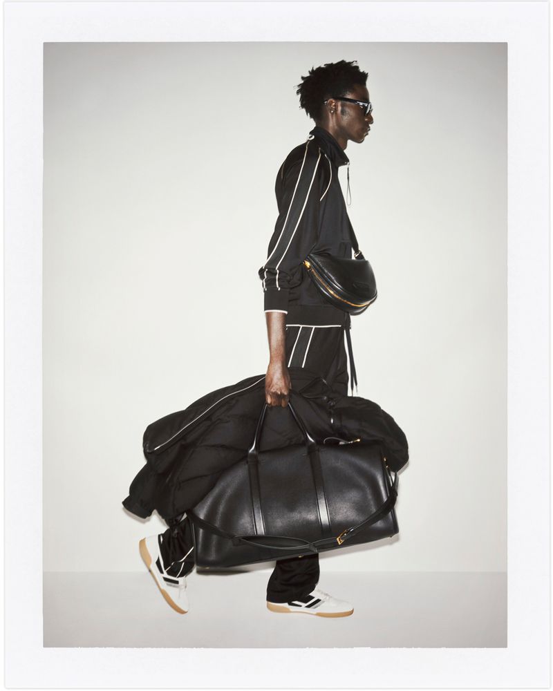 Fashion shot: A Black male model in a tracksuit walks in a studio, carrying a large duffel bag and wearing a black crossbody bag.