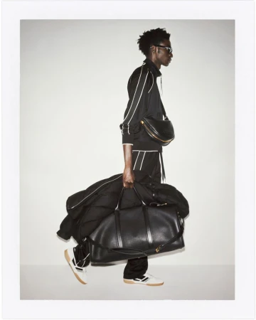 Fashion shot: A Black male model in a tracksuit walks in a studio, carrying a large duffel bag and wearing a black crossbody bag.
