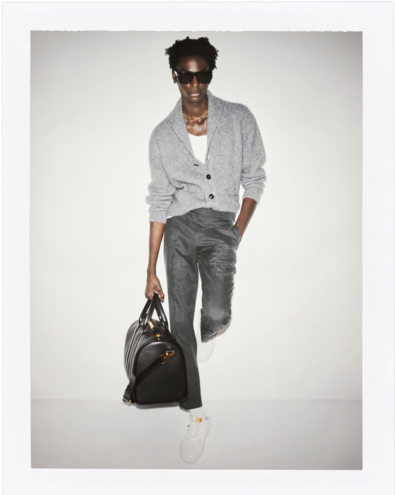 Fashionable adult man wearing sunglasses, knit cardigan, trousers, and sneakers, holding a black leather duffel bag in a studio.