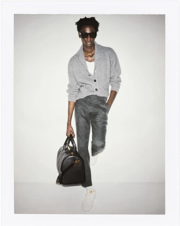 Fashionable adult man wearing sunglasses, knit cardigan, trousers, and sneakers, holding a black leather duffel bag in a studio.